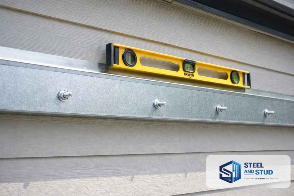 Installing a Steel and Stud galvanized ledger board flush to the house wall with lag screws into structural studs — primary load-transfer attachment for the attached metal carport frame