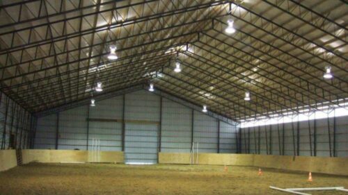 a photo of an indoor horse riding arena. The arena is covered by a large steel frmaed roof with several rows of lights hanging from it. The walls are made of corrugated metal and there are several large windows on the sides.