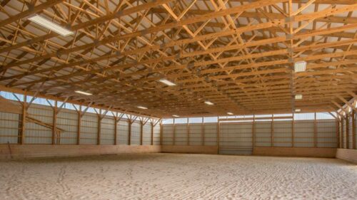 a photo of an indoor horse riding arena. The arena has a sand floor with a pattern of lines. The walls are made of wooden panels and the roof is made of wooden beams.