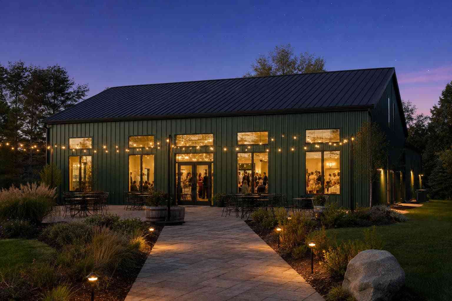 Forest green metal building event venue glowing at dusk with string lights and patio seating