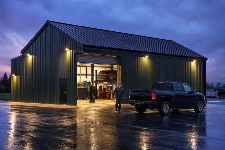 Forest green metal garage at twilight with a black pickup outside the lit lift bay