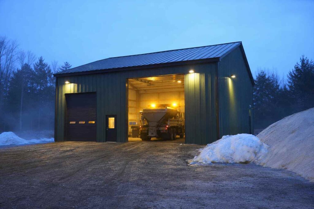 Forest green steel structure at snowy dawn with a salt spreader truck in the lit bay