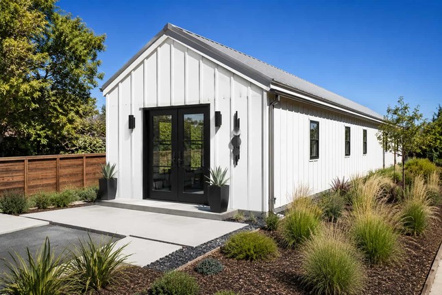 White prefab metal building studio with black double doors and ornamental grass landscaping